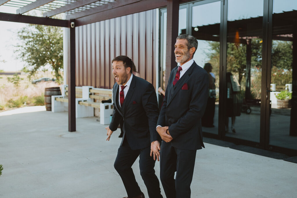 Grooms react with excitement and laughter during a first look with the bridesmaids outside a modern barn-style wedding venue, dressed in matching navy suits and burgundy ties.