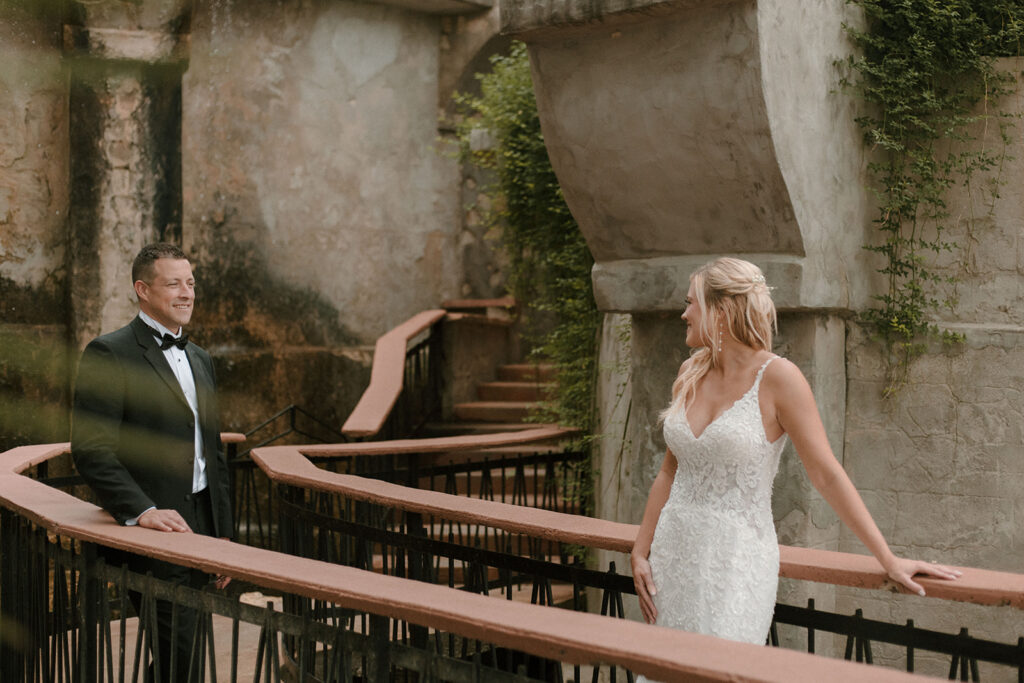 Bride and groom share a romantic first look on a rustic, winding balcony surrounded by textured stone walls and greenery, both smiling as they see each other for the first time.