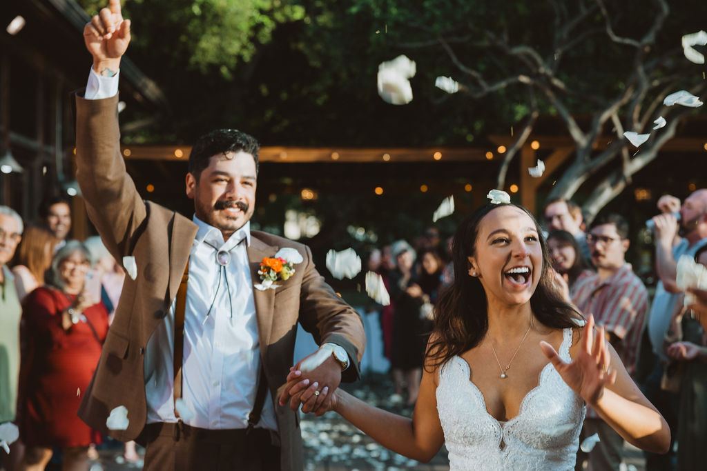A joyful couple celebrates their wedding, with petals being thrown in the air as they walk together, surrounded by smiling guests.