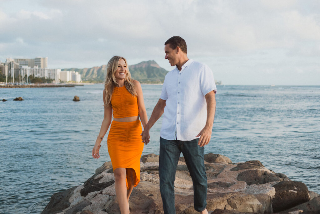A couple walking hand in hand on rocky terrain near the ocean, with a backdrop of a coastal city and mountains in Hawaii. The woman is wearing a bright orange dress, and the man is dressed casually in a white shirt and jeans. destination wedding