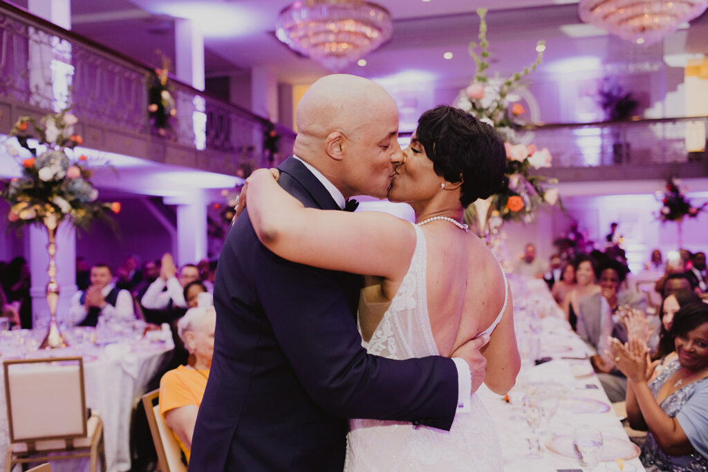 A couple sharing a romantic kiss during their wedding reception, surrounded by elegantly set tables and guests in a beautifully decorated venue.