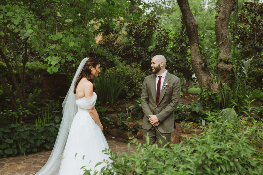 Bride and groom share a joyful first look in a lush garden surrounded by greenery, capturing an emotional and candid moment before the ceremony.