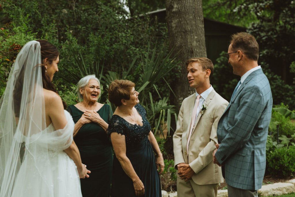 Bride shares an emotional and joyful first look with her family in a lush garden setting, surrounded by parents and grandparents reacting with love and laughter.
