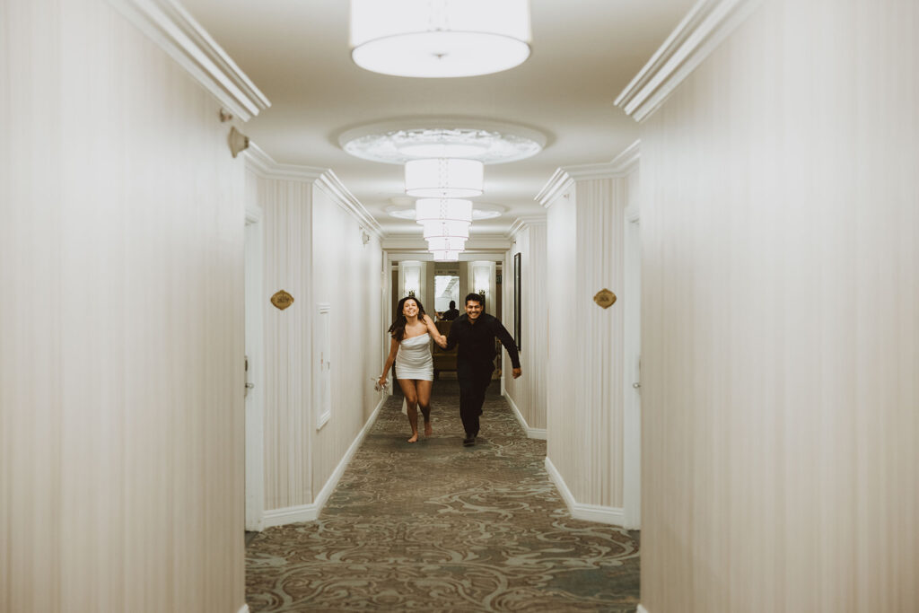 A couple joyfully running down a hotel hallway, with beige wallpaper and stylish lighting fixtures.