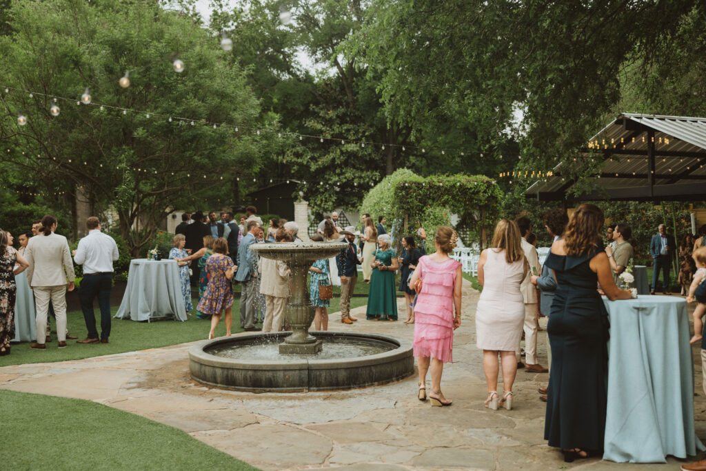 A lively outdoor wedding reception scene with guests mingling around a fountain, dressed in elegant attire surrounded by greenery and decorative lighting.