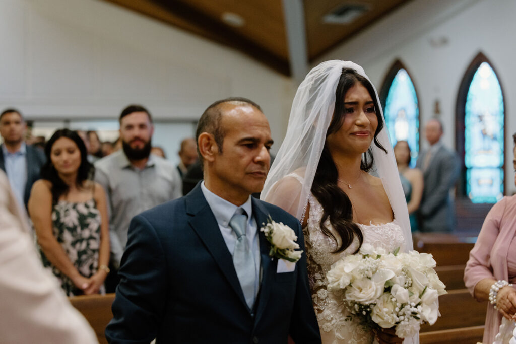 Emotional bride walks down the aisle with her father in a church ceremony, holding a white floral bouquet and fighting back tears as guests look on.