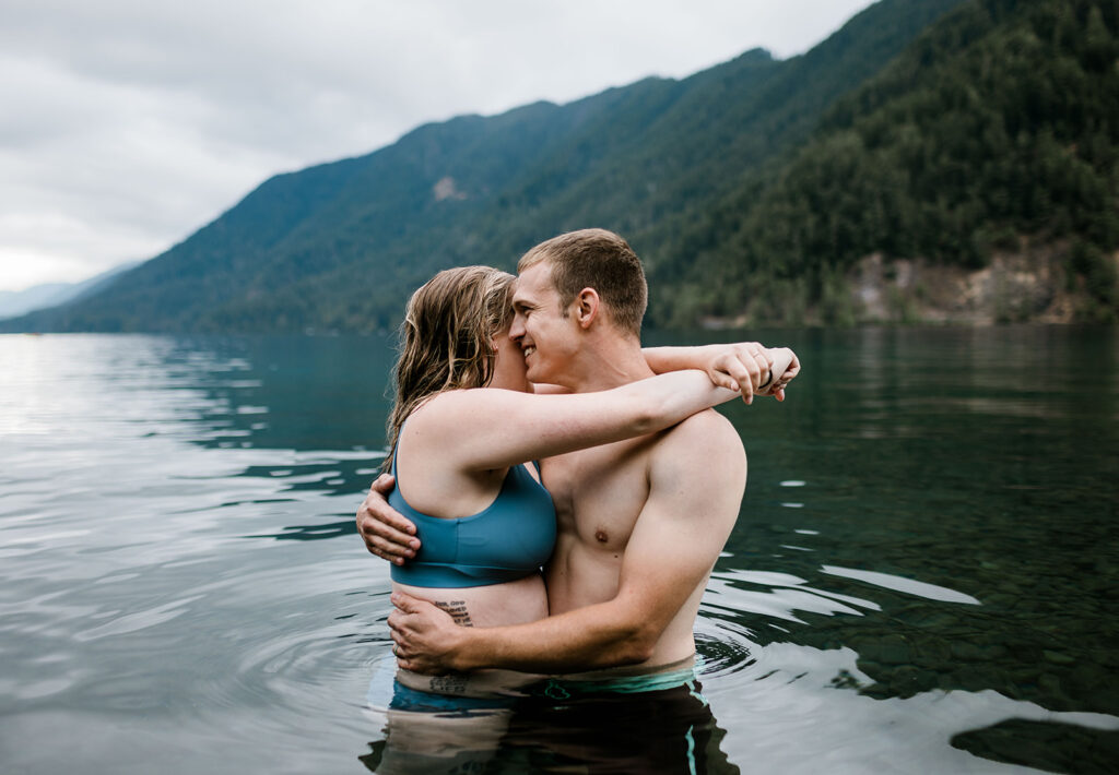 A couple embracing in shallow water with mountains in the background, capturing an intimate moment during their outdoor adventure.