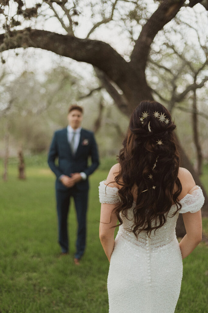 Bride approaches groom for a romantic first look in a quiet, wooded setting, with her long curled hair adorned with celestial hairpins and her off-the-shoulder beaded gown catching the light.