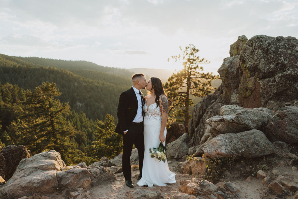 A bride and groom share a kiss on a rocky outcrop during sunset, surrounded by lush green mountains and trees.