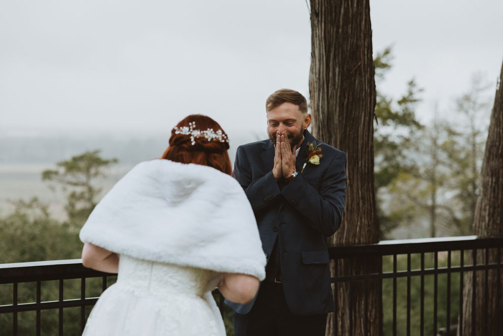 Groom reacts emotionally with hands clasped in awe during a misty first look as the bride approaches in a winter wedding gown and faux fur shawl.