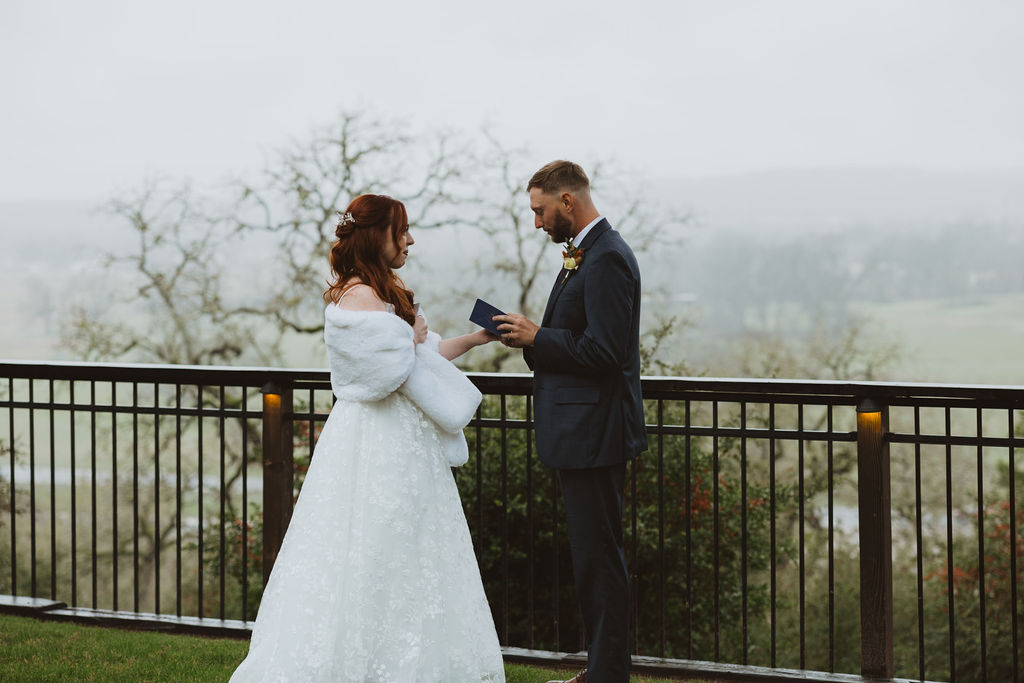 Bride and groom share an emotional private vow exchange during a misty outdoor first look, with the bride in a winter gown and faux fur wrap and the groom reading from a vow book on a scenic overlook.