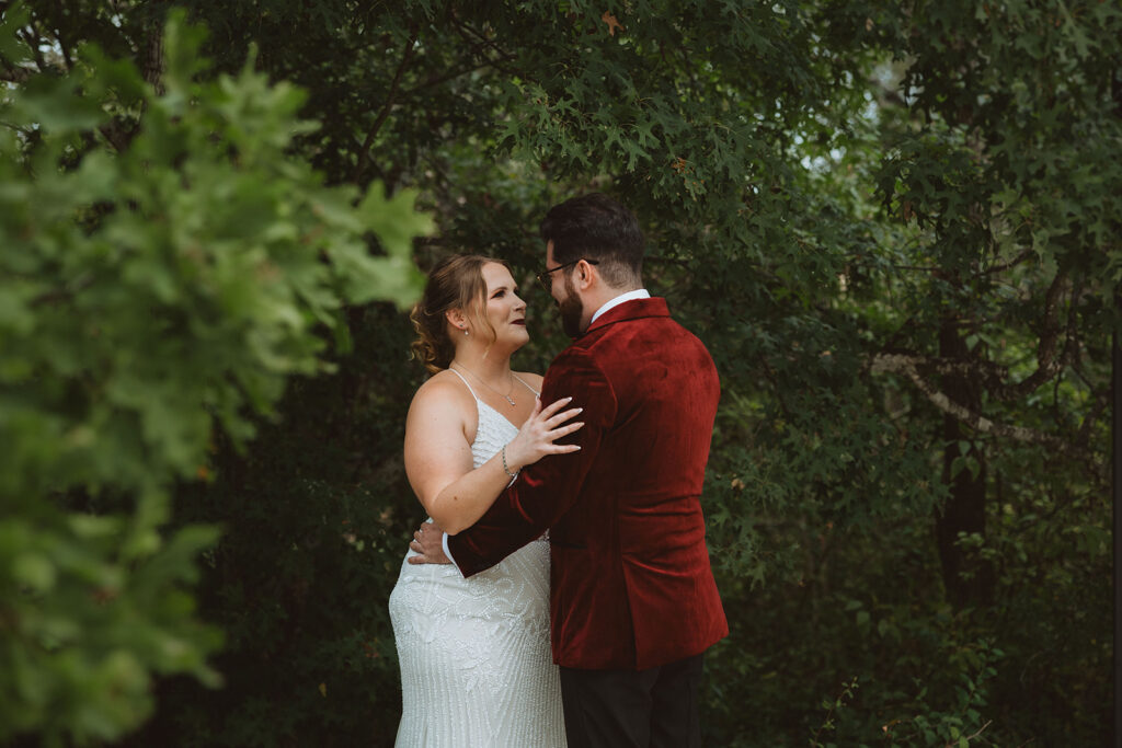 Bride and groom share a heartfelt first look in a wooded outdoor setting, with the bride in a beaded gown and the groom in a bold red velvet jacket, surrounded by lush greenery.
