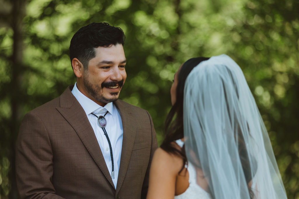 Groom smiles warmly at bride during an outdoor first look, wearing a brown suit and bolo tie as sunlight filters through the trees behind them.