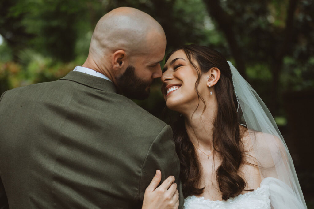 A couple embracing and smiling at each other in a romantic setting, with the groom in a suit and the bride in a wedding dress and veil.