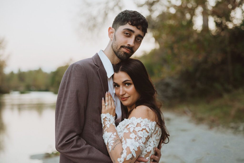 Bride and groom embrace during golden hour by a peaceful lake, with the bride in a floral lace off-the-shoulder gown and the groom in a mauve suit, capturing a quiet, intimate moment.