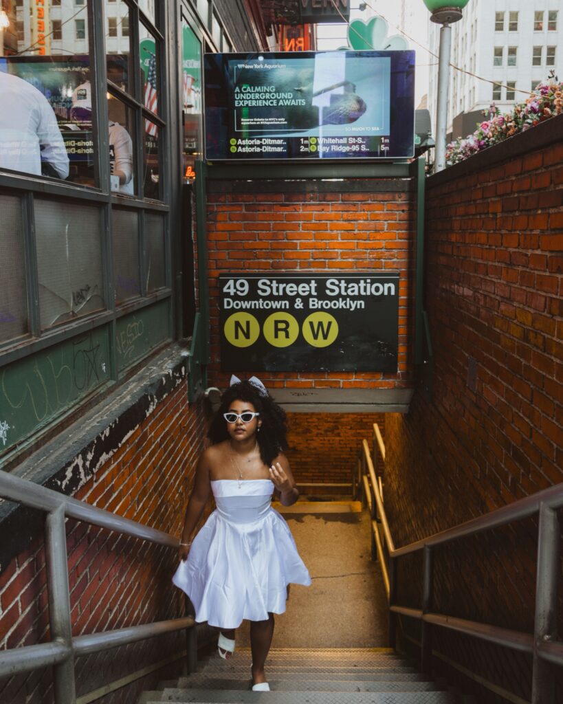 A bride in a white dress and sunglasses walking up the stairs near a subway station in New York City.