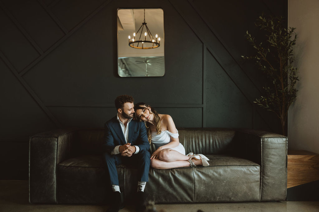 Engaged couple sitting on a modern leather couch against a dark geometric wall, with the woman in a white off-the-shoulder dress resting her head on the man's shoulder. A chandelier is reflected in a mirror above them, adding warmth to the intimate and stylish indoor setting.