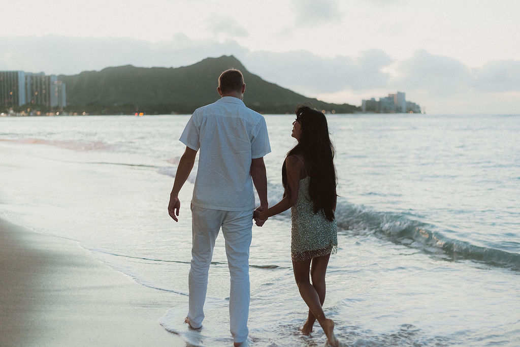 Couple walking barefoot along the shoreline at sunset, holding hands with the ocean on their right and Diamond Head mountain in the background. The man wears an all-white outfit, and the woman wears a sparkly dress, looking up at him with a smile as city lights begin to twinkle in the distance.