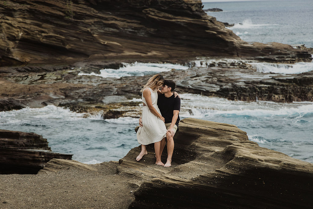 Couple sharing an intimate kiss while sitting barefoot on a rugged rock formation by the ocean. The woman wears a white dress and the man a black shirt, with waves crashing below and dramatic coastal cliffs stretching into the distance behind them.