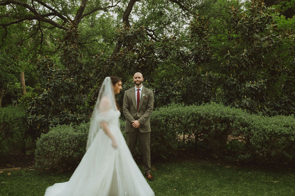Groom stands smiling in anticipation as the bride walks past him during their first look in a lush, green garden surrounded by trees and manicured hedges.