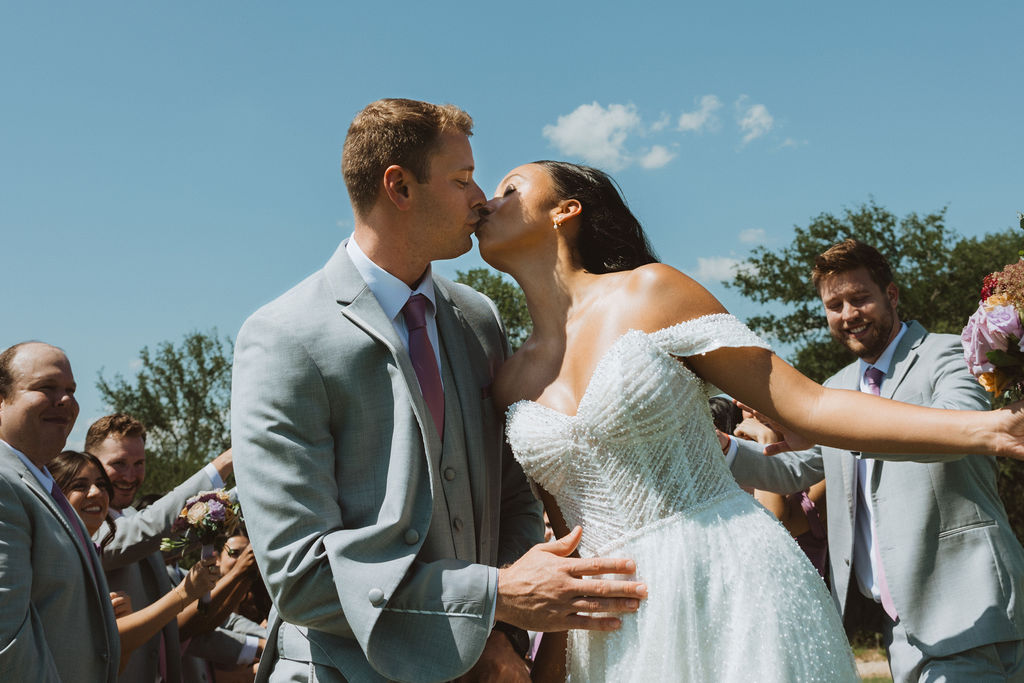 Bride and groom share a kiss under a sunny blue sky, surrounded by their cheering wedding party dressed in light gray suits with lavender accents. The bride wears a sparkling off-the-shoulder gown while the groom holds her waist in a joyful, celebratory moment.