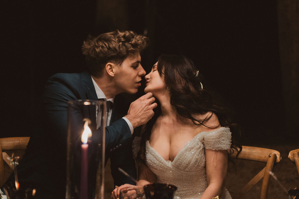 Romantic moment of a groom gently holding his bride’s chin as they lean in for a kiss during their candlelit wedding reception. The bride wears a sparkling off-the-shoulder gown, and the groom is dressed in a dark suit, with a glowing purple candle in the foreground adding to the intimate atmosphere.