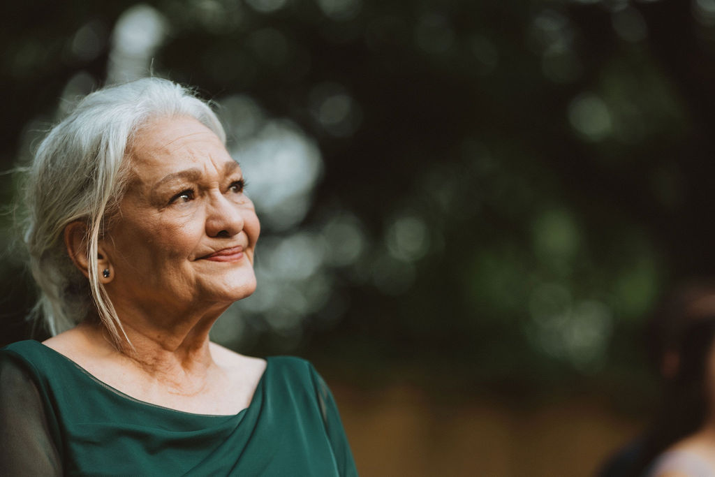 Elderly woman with silver hair styled in a low bun, wearing a deep green dress and small stud earrings, gazes upward with a soft, emotional expression during an outdoor wedding ceremony. The background is softly blurred with natural greenery.