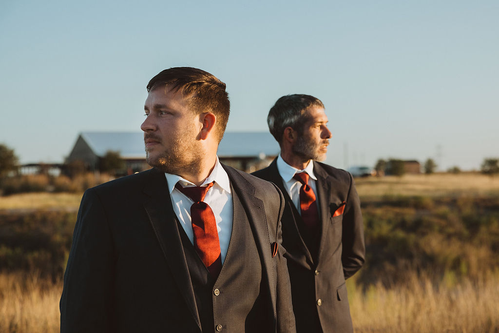 Two grooms in dark suits with burnt orange ties and pocket squares stand in a golden field at sunset, looking in opposite directions. A rustic barn and countryside buildings are visible in the softly blurred background, creating a warm, cinematic atmosphere.