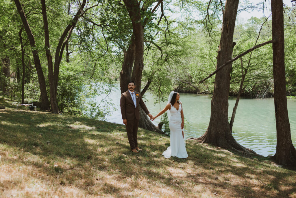 A bride and groom hold hands by the river bank in the Texas Hill Country