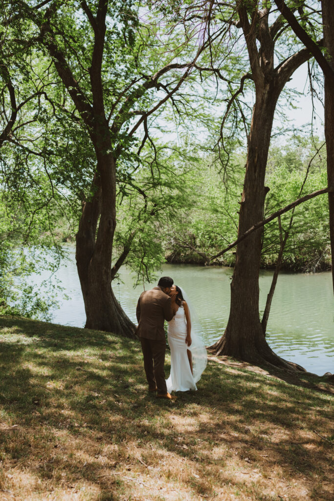 a bride and groom share a kiss by the river bank of the Texas Hill Country