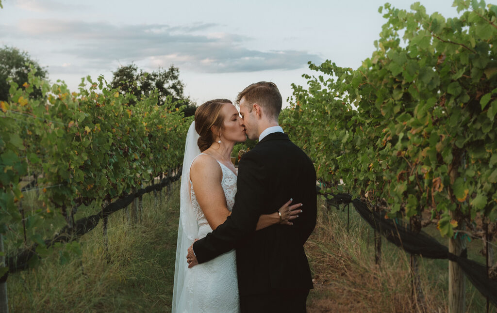 Bride and groom sharing a romantic kiss in the middle of a lush vineyard at sunset in the Texas Hill Country, surrounded by rows of green grapevines.