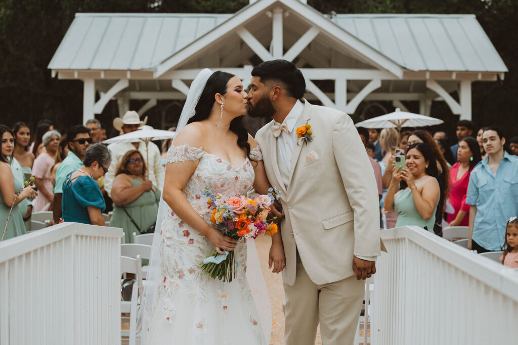Bride and groom share a kiss while walking down the aisle after their ceremony, surrounded by joyful guests and holding a vibrant bouquet of colorful flowers.