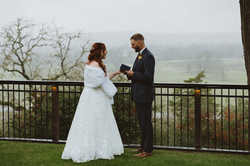 Bride and groom exchange heartfelt vows during an intimate outdoor ceremony on a misty day, standing on a grassy hilltop with fog-covered hills in the background.
