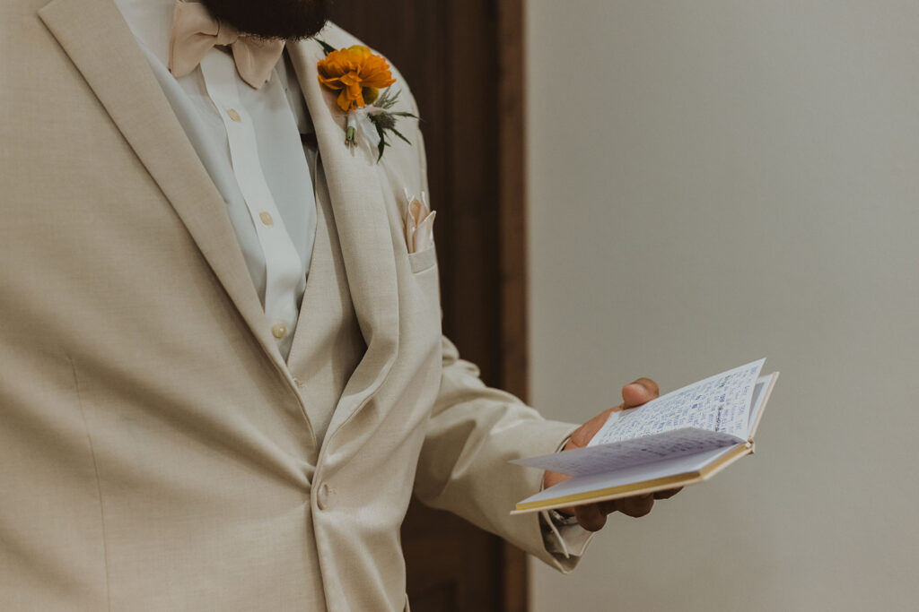 Close-up of groom in a beige suit holding a handwritten vow book, with a yellow boutonniere pinned to his lapel.
