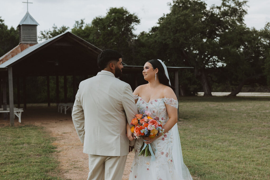 Bride and groom hold hands and smile at each other outside a rustic chapel, with the bride holding a colorful bouquet and wearing a floral lace gown.
