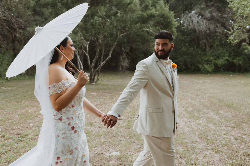 Bride and groom hold hands while walking through a grassy field, with the bride holding a white parasol and smiling, and the groom looking back at her with a joyful expression.