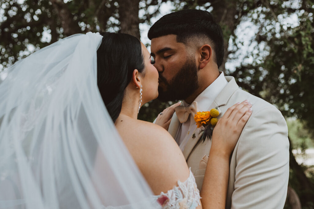 Bride and groom share an intimate kiss outdoors beneath trees, with the bride wearing a long veil and the groom in a cream suit and bow tie adorned with a yellow boutonniere.