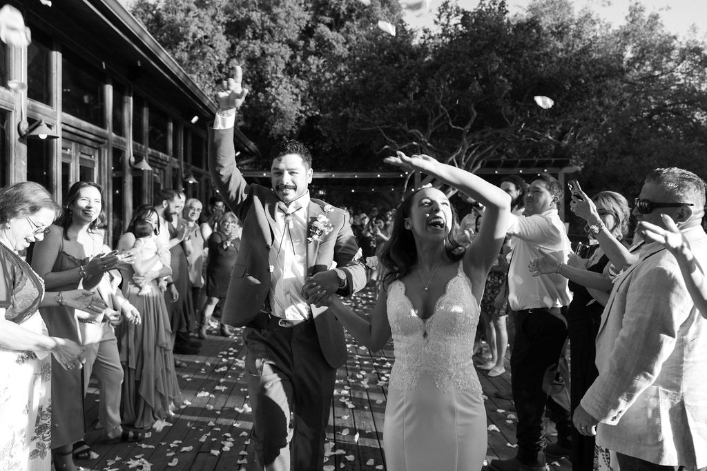 lack and white photo of a joyful bride and groom walking hand-in-hand through a crowd of cheering guests throwing flower petals during their wedding exit.  