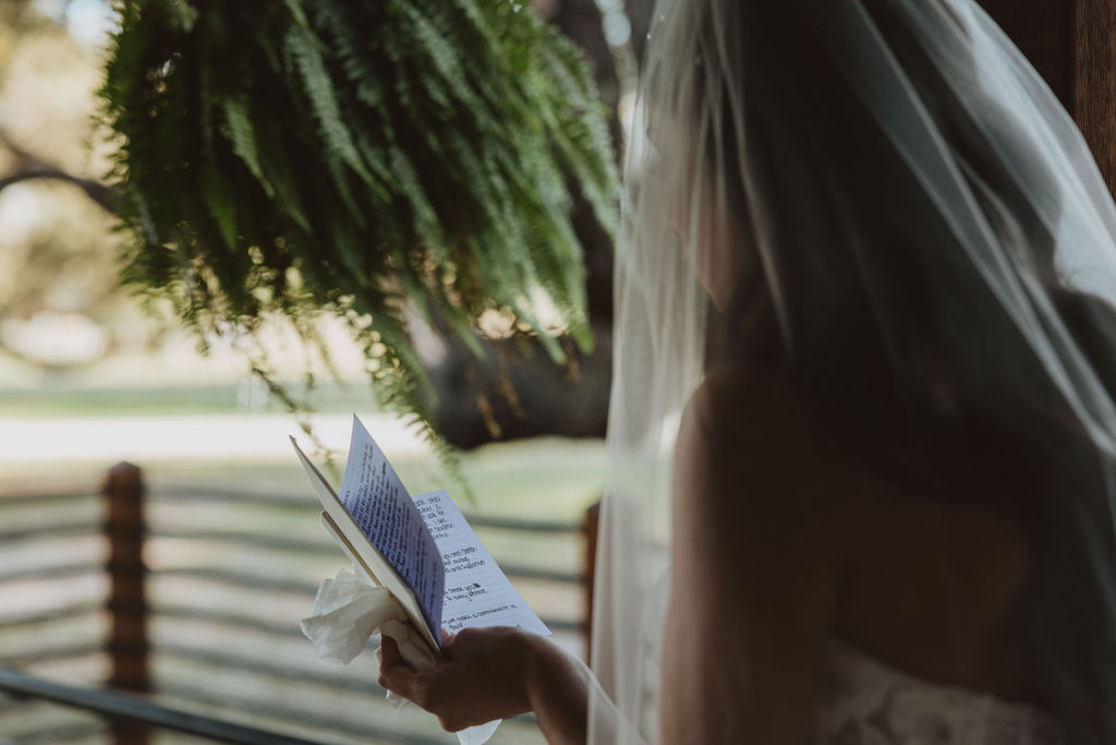 Bride stands quietly in soft natural light, holding a handwritten letter and tissue, reading heartfelt vows before the ceremony.