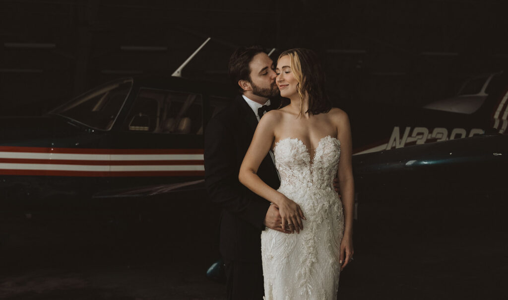 Groom embraces and kisses his bride as they stand in front of a vintage airplane inside a dimly lit hangar, the bride glowing in an intricately beaded strapless gown.