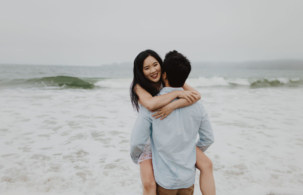 Smiling woman wraps her arms around her partner's shoulders as he lifts her up at the edge of the ocean, with gentle waves and a foggy coastline in the background.