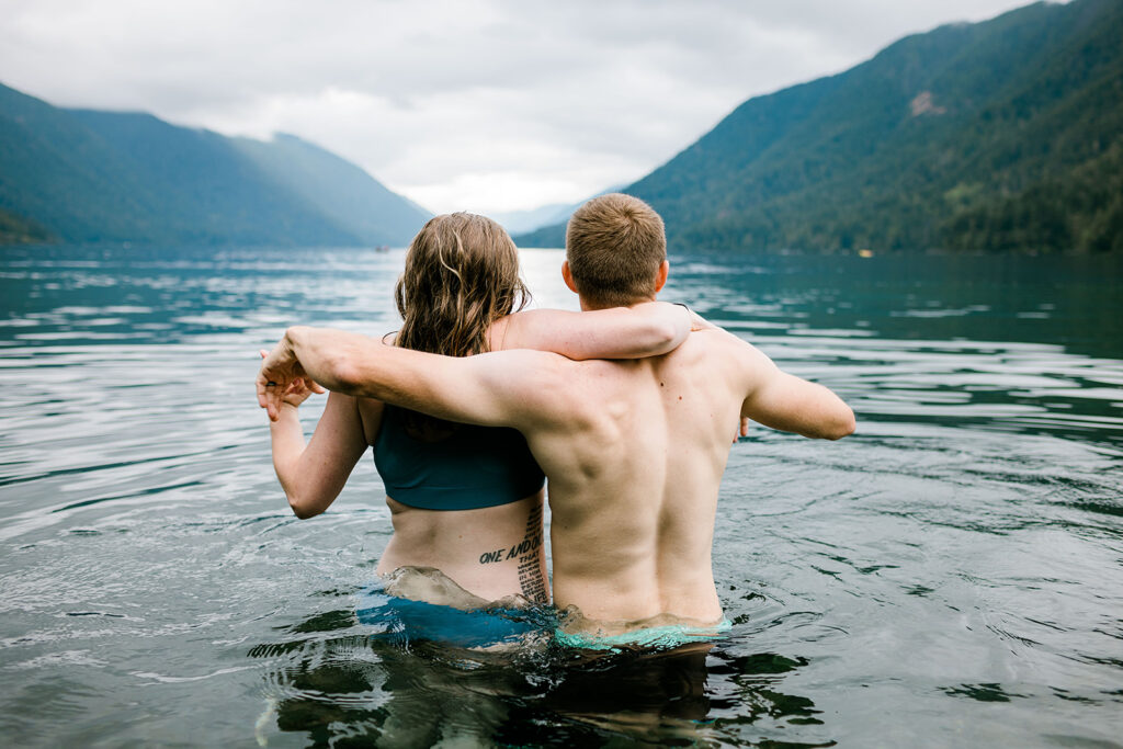 Couple stands waist-deep in a serene mountain lake, arms wrapped around each other, looking out at the misty forest-covered peaks in the distance.