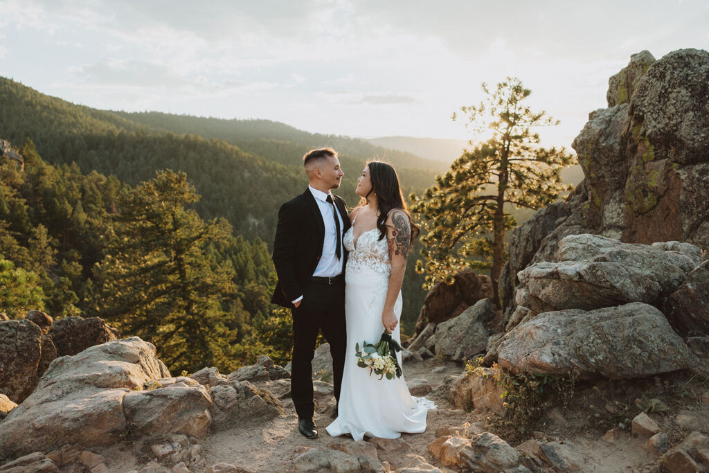 Bride and groom stand on a rocky mountain overlook at sunset, surrounded by pine trees and golden light, sharing an intimate moment during their outdoor wedding in Boulder, Colorado.