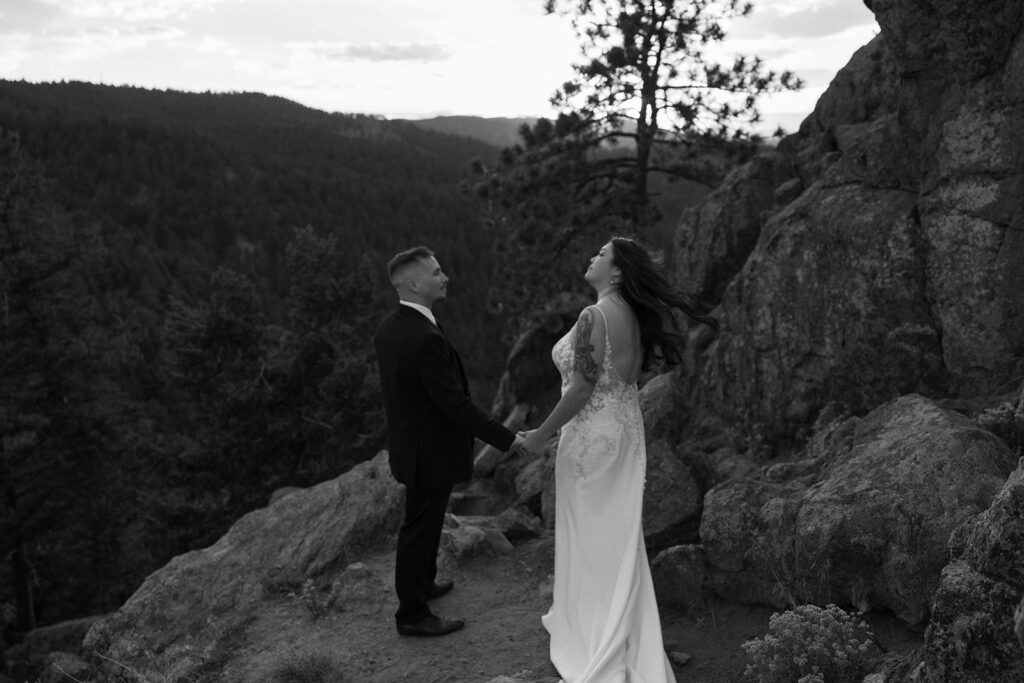 Black and white photo of a bride and groom holding hands on a rocky cliffside, surrounded by forested mountains at sunset, as the bride laughs with her hair blowing in the wind.
