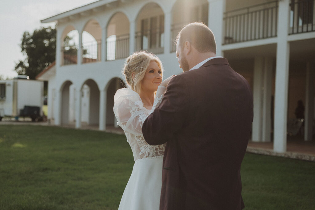 Bride and groom share a quiet, intimate moment in front of a white Mediterranean-style venue, bathed in golden sunlight with lush green grass surrounding them.