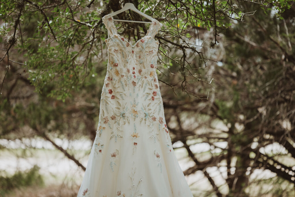 Floral embroidered wedding dress hanging from a tree branch in a wooded outdoor setting, surrounded by natural greenery.