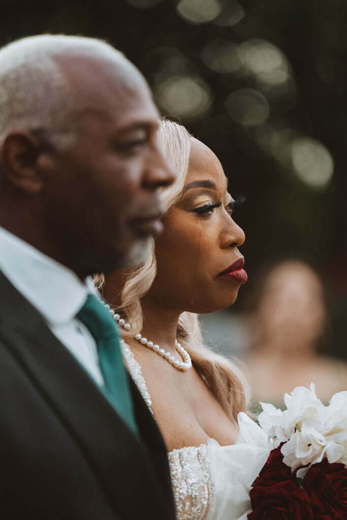  Bride walks down the aisle with tears streaming down her face, holding a bouquet of white and red flowers, accompanied by her father during an emotional wedding moment.