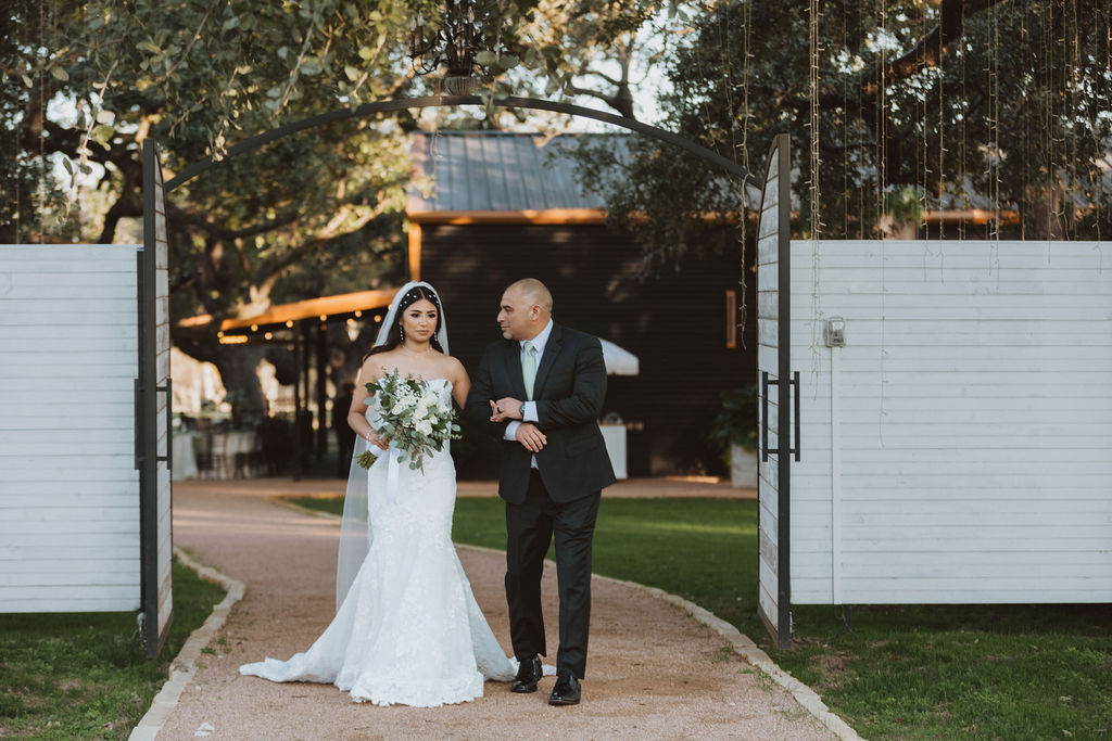 Bride in a strapless lace gown walks arm-in-arm with her escort through open gates toward the ceremony, holding a lush bouquet of greenery and white flowers.