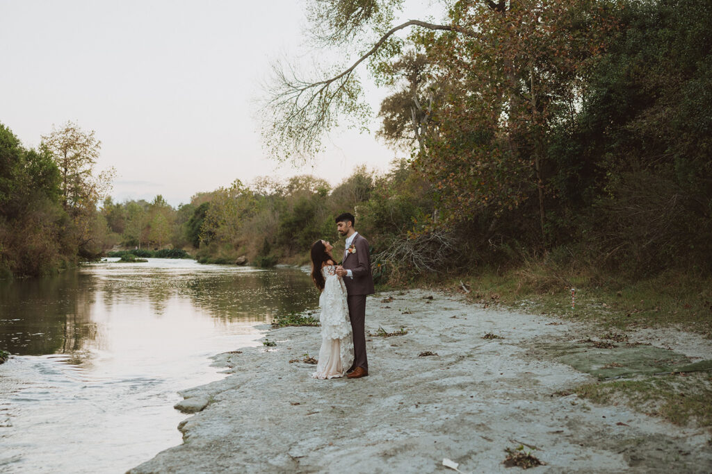 Bride and groom standing together on a quiet riverbank in the Texas Hill Country at sunset, surrounded by trees with soft autumn foliage and serene water.