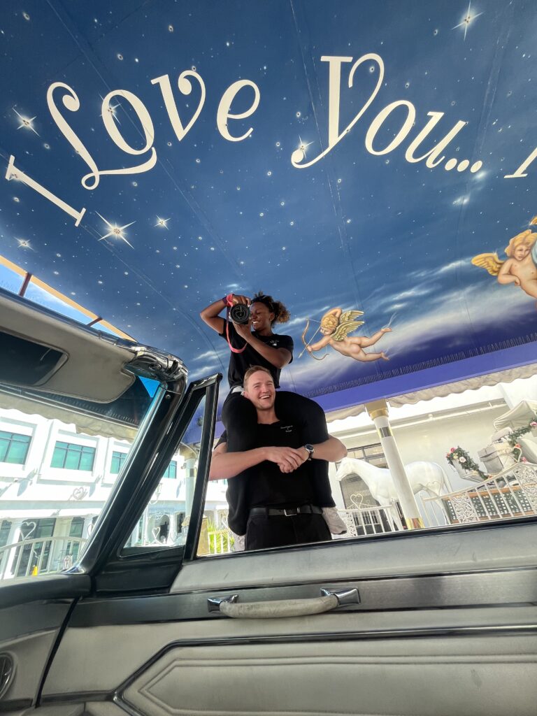 Behind-the-scenes moment of a joyful photographer standing on a teammate’s shoulders to capture a creative shot, under a whimsical “I Love You” sign at a wedding venue decorated with cherubs and a classic car.